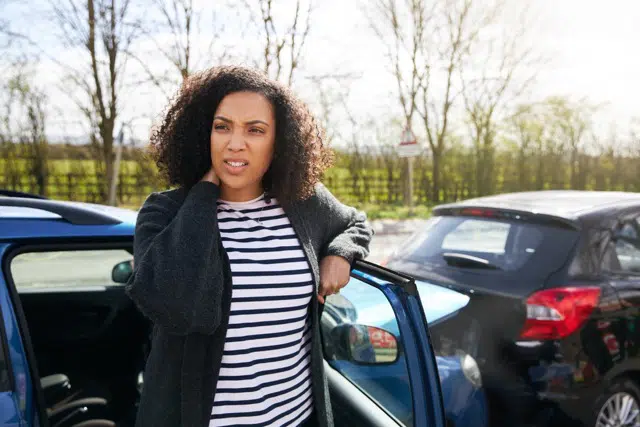 Young woman rubbing neck in pain from whiplash injury standing by damaged car after traffic accident