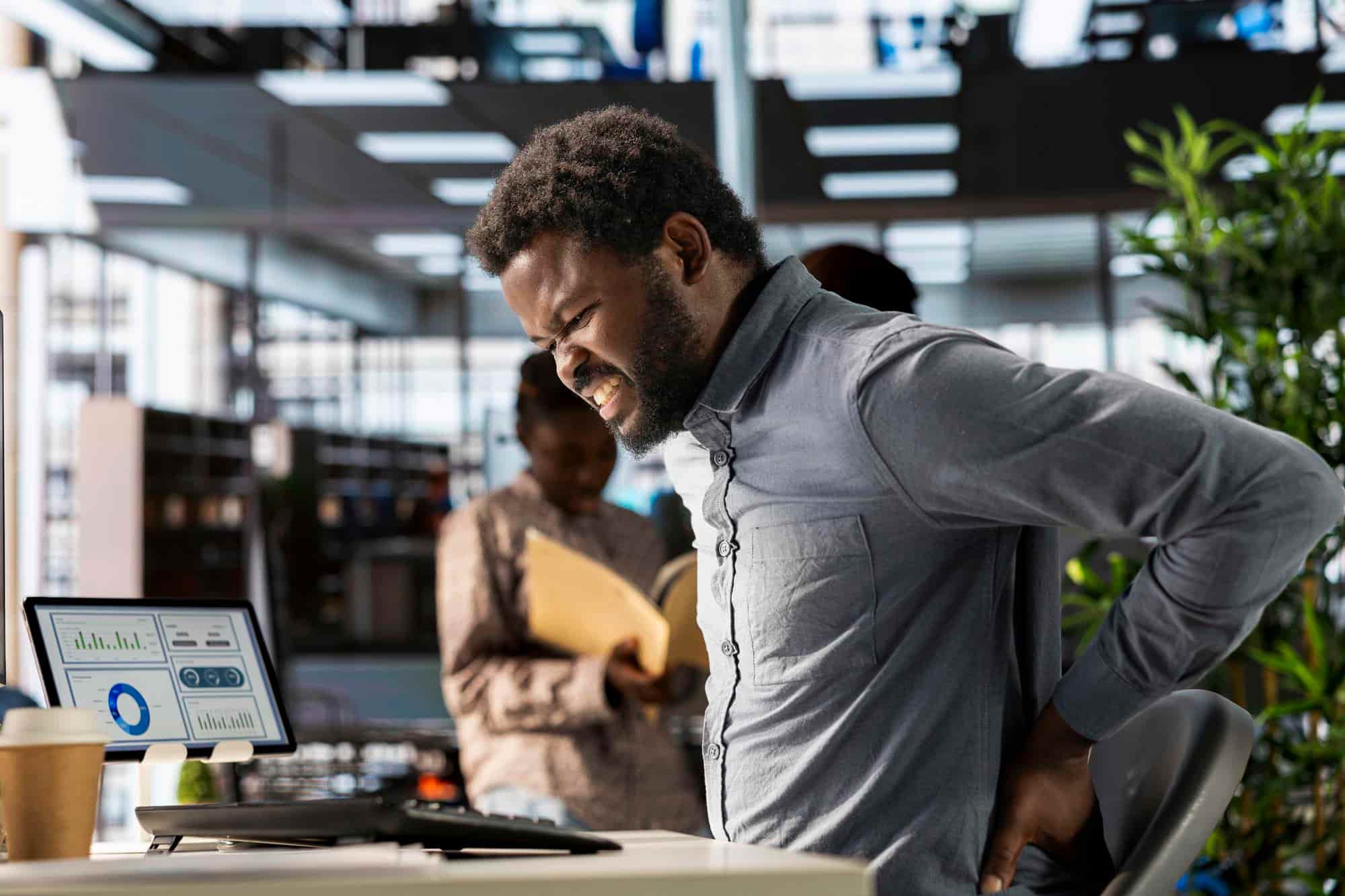 A man in a grey shirt wincing in pain and clutching his lower back while sitting in an office chair, illustrating how back pain interferes with daily work tasks.