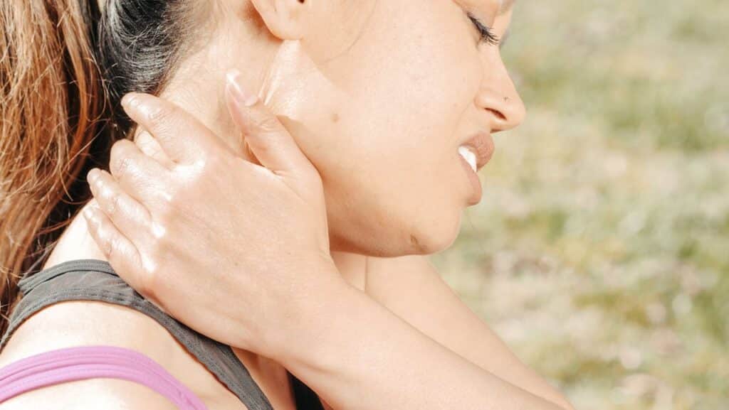 Close-up of a person in athletic wear wincing and clutching their neck in sharp pain outdoors, illustrating severe neck stiffness and movement restrictions.