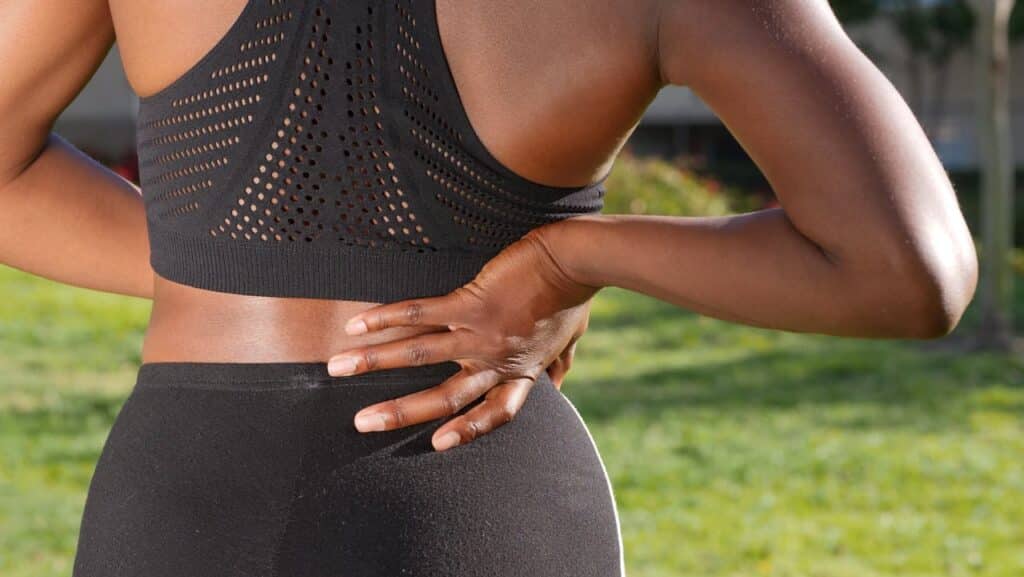 A person in athletic wear holding their lower back in pain while standing outdoors in a park.