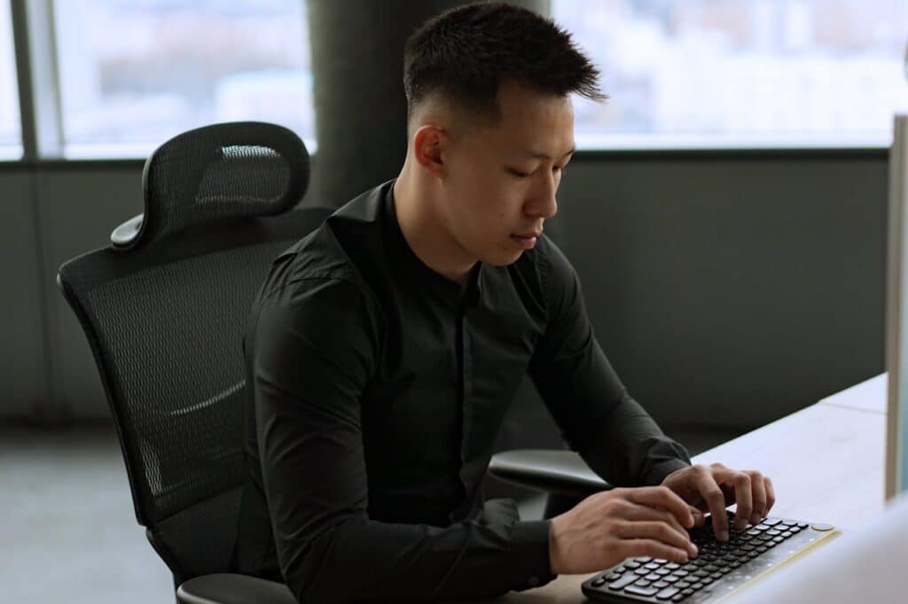 A professional man sitting at a desk with an ergonomic chair, maintaining an upright posture while typing on a keyboard.