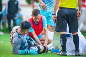 A soccer goalkeeper lies injured on the turf field while a teammate kneels to check on his leg and a referee and medical trainer stand by to assist.