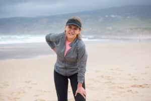 An older woman in athletic wear stands on a beach with a pained expression, clutching her lower back and resting her hand on her knee.