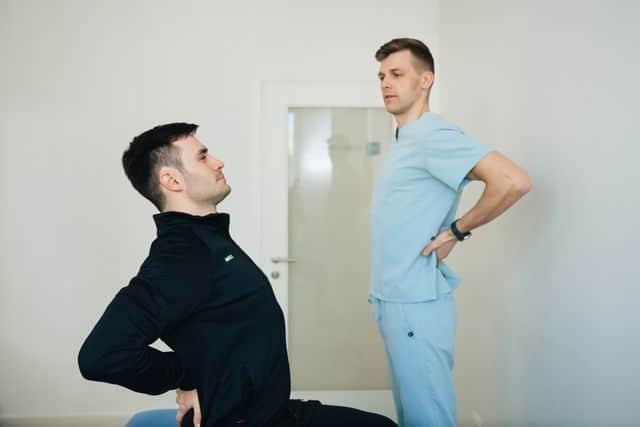 A male patient in athletic wear performing a back-bending extension test while a clinician in light blue scrubs observes his posture and range of motion.