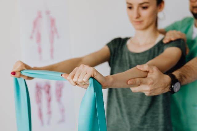 A female patient pulling a green resistance band while a practitioner in scrubs stabilizes her forearm and shoulder posture.