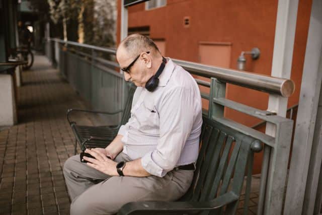 A man sitting on an outdoor metal bench, slouched forward with his head tilted down while using a mobile smartphone.