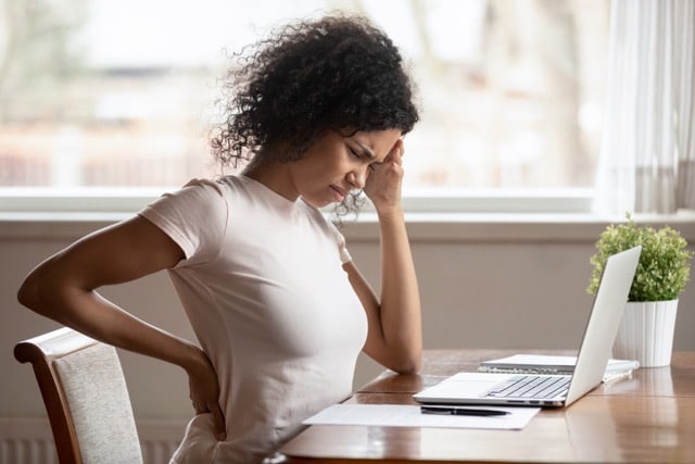 A woman sitting at a laptop on a wooden desk, grimacing in discomfort while clutching her lower back and forehead.
