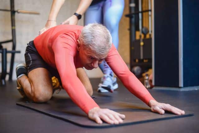 A physical therapist supervising a man doing a kneeling lumbar stretch on an exercise mat in a clinic.